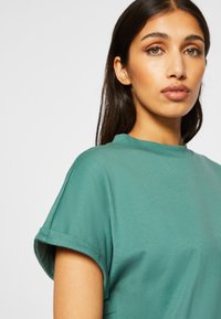 Teal bomullstop med et minimalistisk design, korte ermer og høy hals. Myk tekstur, med rullede ermer og en avslappet passform.