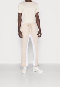 Beige t-shirt med rund hals, tillsammans med beige byxor som har vita sidopaneler, elastisk midja och dragskojustering. Vita sneakers.