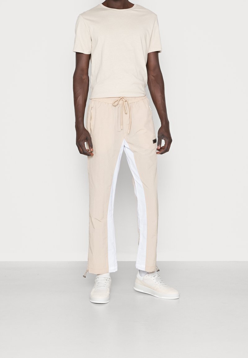 Beige t-shirt med rund hals, tillsammans med beige byxor som har vita sidopaneler, elastisk midja och dragskojustering. Vita sneakers.