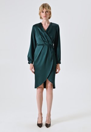 Dunkelgrünes Satin-Wickeldress mit langen Ärmeln, tiefem V-Ausschnitt, Taillenschnürung und asymmetrischem Saum. Kombiniert mit schwarzen spitzen Absatzschuhen.