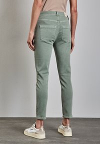 Jeans de mezclilla verdes con un corte ajustado, cinco bolsillos y detalle en el yugo trasero. Llevados con zapatillas blancas con un acento metálico.