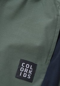 Grön tyg med synlig sömnad och en svart fyrkantig lapp med texten "COLOR KIDS" i vita versaler.