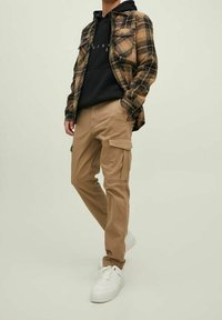 Persona che indossa pantaloni cargo beige, sneakers bianche, felpa nera con cappuccio e camicia oversize a quadri marroni su uno sfondo chiaro e semplice.