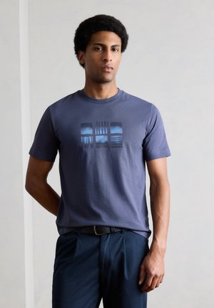 Άνδρας με κοντά σγουρά μαλλιά, φορώντας μπλε μπλουζάκι με γραφικά και σκούρο μπλε παντελόνι, μπροστά σε απλό ανοιχτό γκρί φόντο.