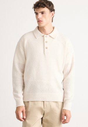 Ung man iklädd en krämfärgad långärmad polo-tröja med tre knappar och beige byxor, stående mot en enkel vit bakgrund.