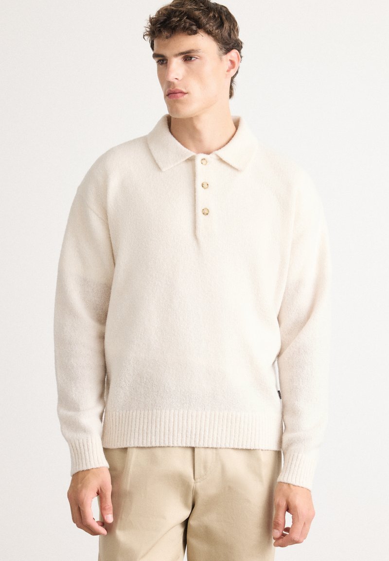 Joven vestido con un suéter de polo de manga larga color crema, con tres botones, y pantalones beiges, de pie frente a un fondo blanco liso.