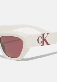 Baltos plastikinės akiniai nuo saulės su kampuota forma, bordinės spalvos objektyvai ir raudonas "cK" logotipas šone. Lygi tekstūra, drąsi spalvų kontrastas.