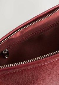 Interior de bolso de cuero burdeos con un forro suave de satén, que cuenta con un zipper plateado y herrajes metálicos. Detalle de superficie texturizada visible.