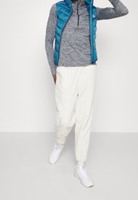 Padded teal Weste über einem grauen Langarmshirt, kombiniert mit weißen, tapered Jogginghosen und weißen Sneakern mit pinken Akzenten.