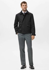 Chaqueta negra de botones con dos bolsillos frontales, llevada sobre una camisa blanca y corbata; combinada con pantalones grises y zapatos negros.