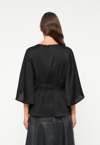 Zwarte blouse met wijde mouwen, geplooide taille, zachte textuur en rits aan de achterkant. G gedragen over een zwarte leren rok.