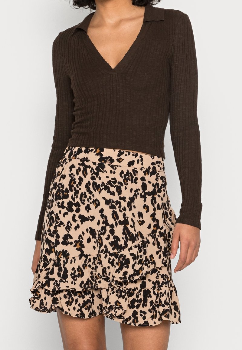 Femme portant un haut à manches longues côtelé marron foncé avec col en V et une jupe beige avec imprimé animal noir et marron et des détails à volants.