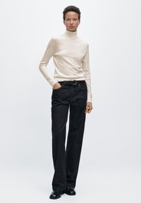 Beige coltrui van zacht breisel, gecombineerd met donker denim, wijde broek met riemlussen en zijzakken. Zwarte schoenen.