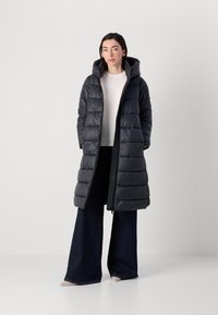 Een vrouw staat in een zwarte pufferjas met een capuchon, gecombineerd met een lichte trui en flared donkere jeans, tegen een eenvoudige achtergrond.