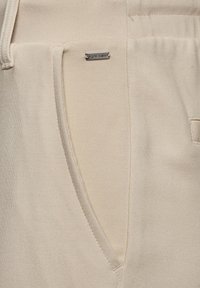 Beige joggerbroeken van een gladde stof, met een zijzak en een klein metalen merkteken. Het ontwerp bevat subtiele naden en een relaxte pasvorm.