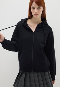 Sudadera negra con cremallera, capucha ajustable y bolsillos delanteros, hecha de material suave. Llevada con una falda plisada a cuadros gris.