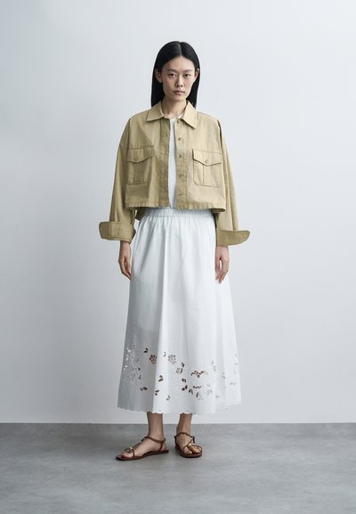 Γυναίκα στέκεται σε απλό φόντο, φοράει χακί κοντό μπουφάν, λευκή κεντημένη midi φούστα και καφέ επίπεδα σανδάλια.