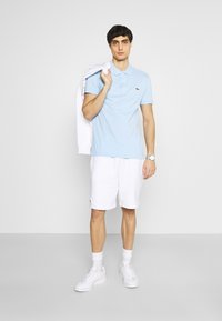 Polo azzurro chiaro con colletto, abbinato a pantaloni corti bianchi. Scarpe da ginnastica bianche e un maglione bianco gettato su una spalla. Logo Lacoste visibile.
