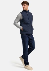 Chaleco acolchado azul marino con un cuello alto, dos bolsillos frontales y cierres de presión, usado sobre una camiseta de manga larga gris claro y jeans oscuros.