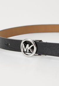 Curea din piele neagră cu o cataramă circulară argintie pe care sunt inscripționate literele "MK", având o suprafață texturată și un design minimalist.