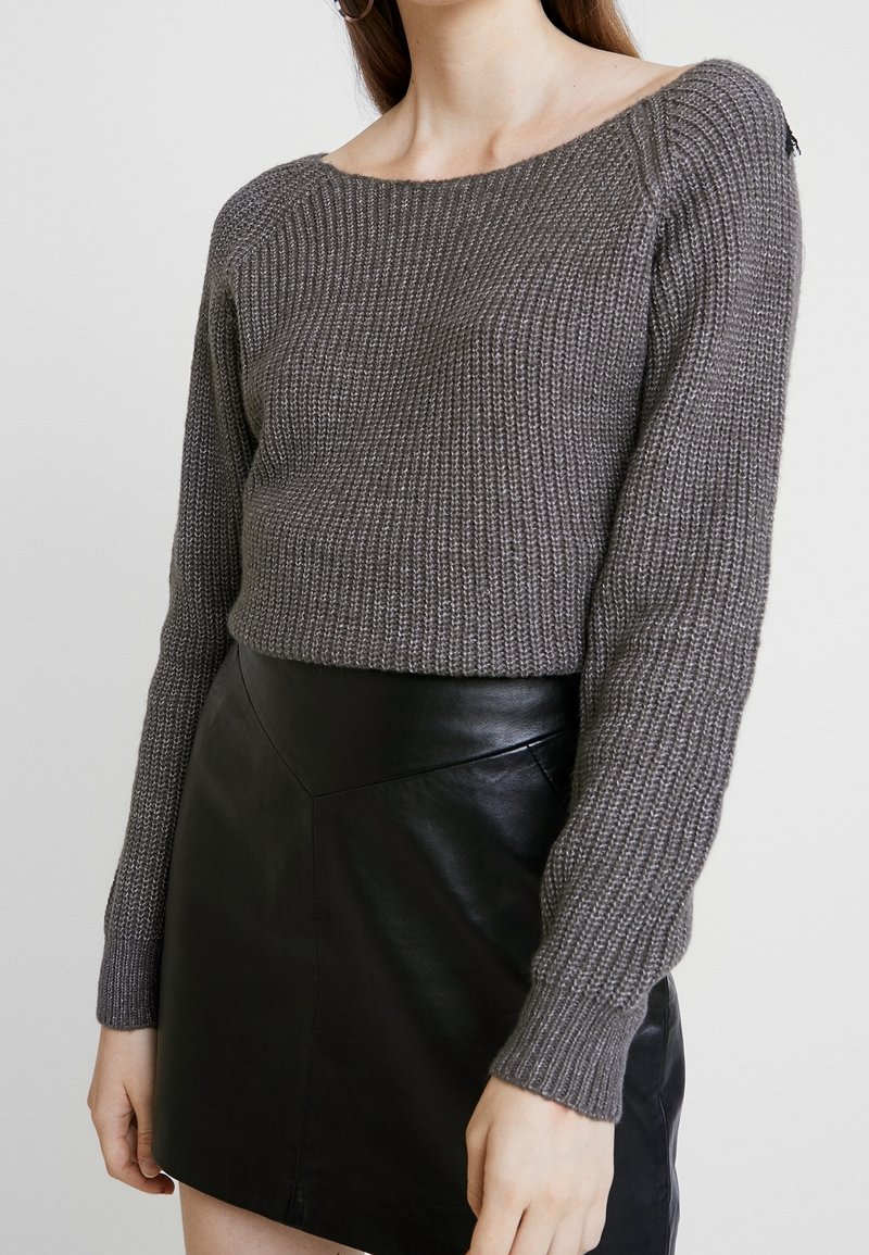 Femme portant un pull en maille gris à manches longues et une mini jupe en cuir noire devant un fond clair uni.