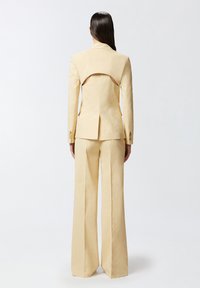 Completo beige su misura con giacca aderente caratterizzata da dettagli cut-out sulla schiena e pantaloni a gamba ampia. Realizzato in un tessuto morbido e leggero.