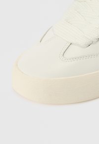 Λευκό δερμάτινο αθλητικό παπούτσι με υφασμένη λεπτομέρεια στο κορδόνι και ανάγλυφη ελαστική σόλα σε ανοιχτό εκρού, με αεροδυναμικό σχέδιο και διακριτικές ραφές.