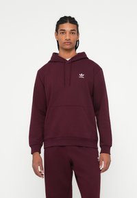 Fiatal férfi, akinek fonott haja van, bordó Adidas kapucnis pulóvert és illő melegítőt visel, egy sima fehér háttér előtt áll.