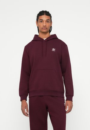 Mladý muž s opletenými vlasmi, oblečený v bordovom mikinovom svetríku Adidas a zodpovedajúcich teplákach, postávajúci pred jednoduchým bielym pozadím.