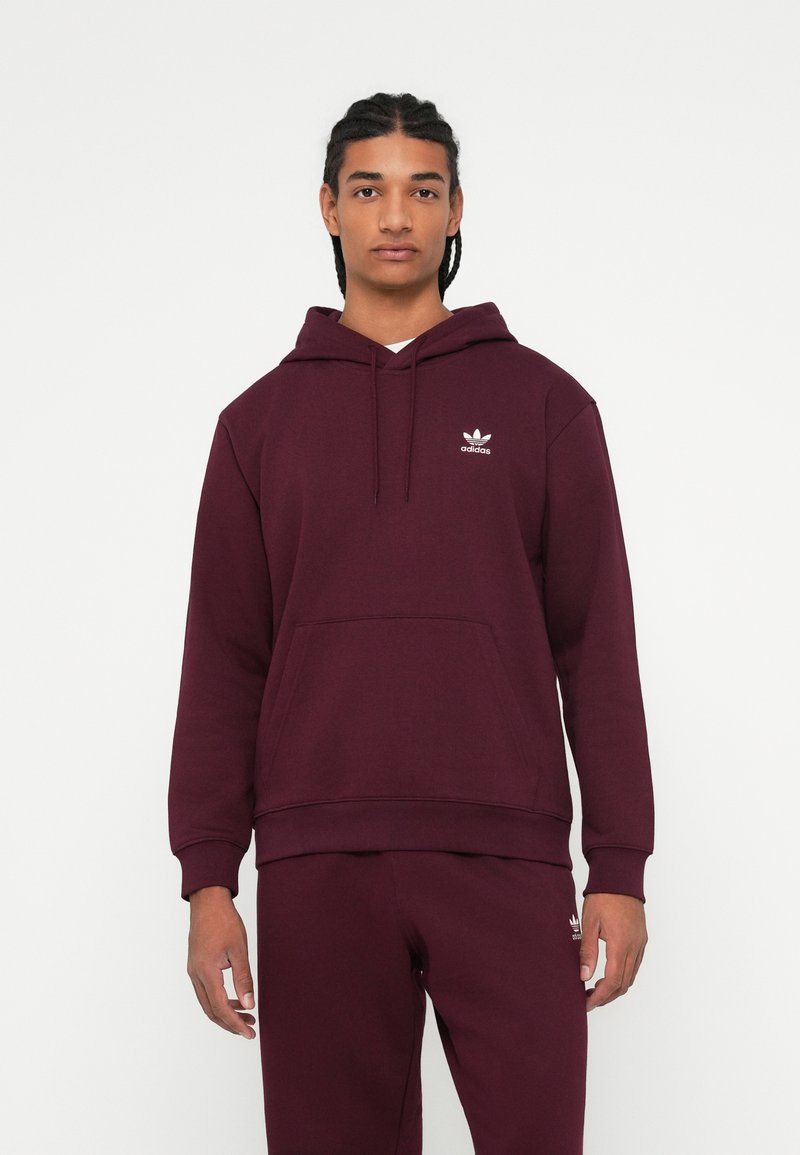 Fiatal férfi, akinek fonott haja van, bordó Adidas kapucnis pulóvert és illő melegítőt visel, egy sima fehér háttér előtt áll.