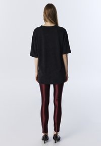 Przesadzony czarny bawełniany t-shirt z krótkimi rękawami, połączony z błyszczącymi bordowymi legginsami i szpiczastymi czarnymi szpilkami, widok z tyłu.