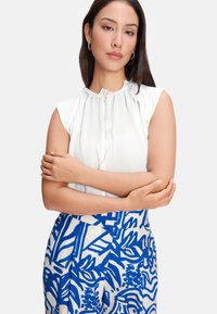 White sleeveless blouse with ruffled neckline and button detail, paired with blue and cream patterned skirt featuring tropical motifs.