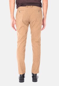 Uomo in pantaloni di velluto a coste beige, scarpe nere e camicia scura, in piedi di spalle contro uno sfondo bianco semplice.