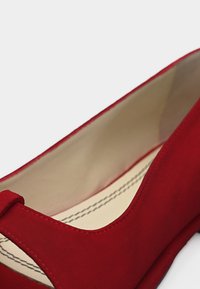 Κόκκινο σουέντ επίπεδο παπούτσι με στρογγυλή μύτη και μπεζ εσωτερική επένδυση, με ορατή ραφή στην εσωτερική σόλα.
