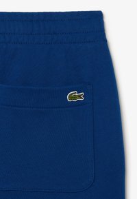 Blaue Baumwollshorts mit einem elastischen Bund, ausgestattet mit einer einzelnen Gesäßtasche und einem grünen, gestickten Krokodil-Logo. Glatte Textur.