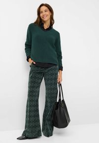 Donkergroene gebreide trui met een V-hals, gecombineerd met groene wijde broek met patroon en een gestructureerd design, en een zwarte handtas.