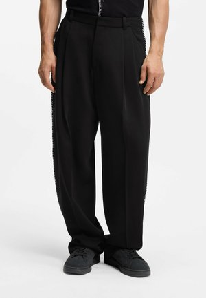 Mann trägt schwarze, plissierte, weit geschnittene Hosen und schwarze Sneaker, steht vor einem weißen Hintergrund.