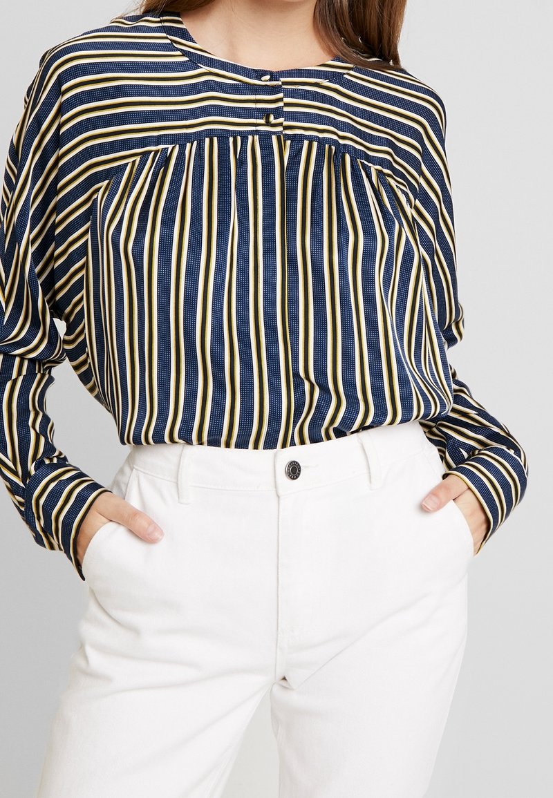 Blouse rayée bleu marine et blanche avec accents jaunes, encolure boutonnée, taille froncée et manches longues, associée à un pantalon blanc.