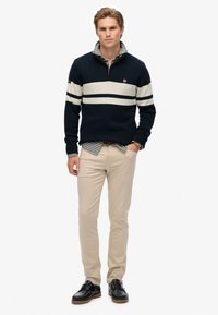 Joven de pie con una mano en el bolsillo, vistiendo un suéter a rayas azul marino sobre una camisa de cuadros, pantalones beige y zapatos negros sobre un fondo blanco.