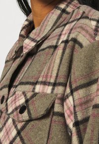 Camisa de cuadros en verde oliva, rosa y negro. Hecha de tela texturizada con dos bolsillos delanteros con botones negros y un collar.