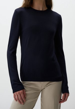 Femme portant un pull noir ajusté à manches longues et un pantalon beige, debout devant un fond clair uni.