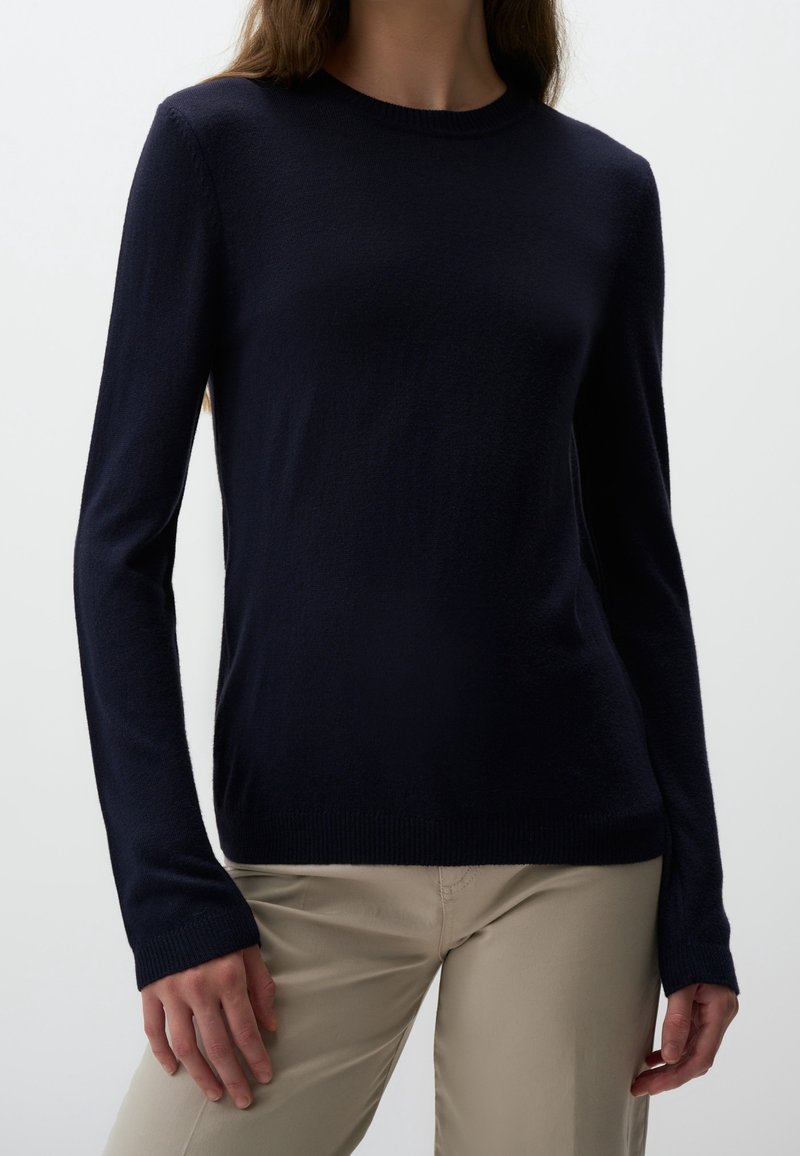 Femme portant un pull noir ajusté à manches longues et un pantalon beige, debout devant un fond clair uni.