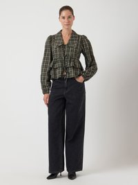 Žena stoji odjevena u zelenu kariranu bluzu dugih rukava s vezicama sprijeda, crne široke traperice i crne cipele s oštrim vrhom, na neutralnoj pozadini.