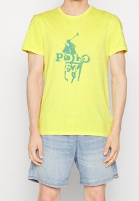 Keltainen lyhythihainen T-paita pyöreällä kaula-aukolla, jossa on sininen poloauran graafinen kuva ja teksti "POLO 67." Puuvillamateriaali, jossa on tekstuuripinta.
