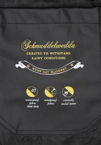 Černá látka s zlatým textem a symboly. Uvádí vlastnosti: voděodolnost (3000 mm), větruodolnost, kriticky utěsněné švy. Text ve dvou jazycích.