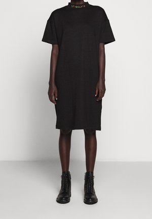 Persona con vestido negro de manga corta hasta la rodilla con "STIEGLITZ" en letras de colores en el cuello y botas negras hasta el tobillo con cordones.