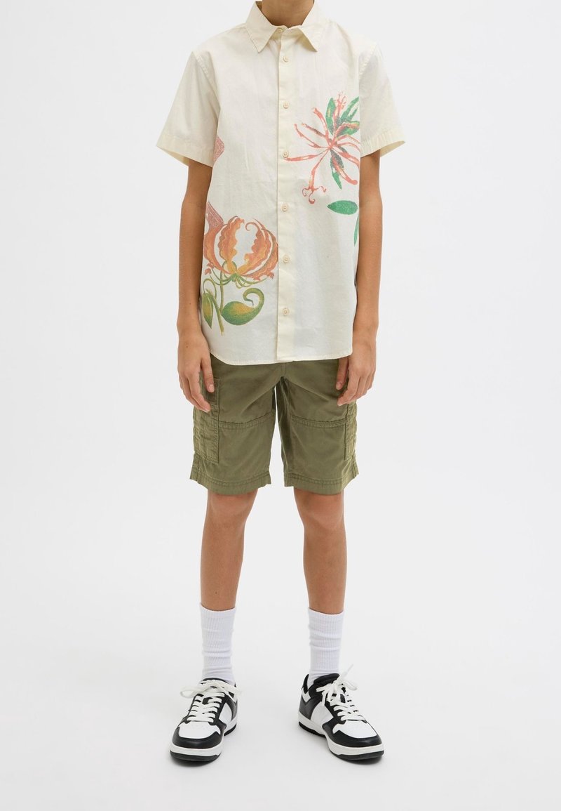 Garçon portant une chemise beige à manches courtes avec des imprimés floraux, un short cargo vert olive, des chaussettes blanches et des baskets noires et blanches.