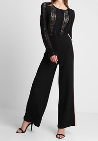 Schwarzes Spitzenoberteil mit langen Ärmeln und vertikalen Streifen, kombiniert mit hoch taillierter schwarzer Hose mit weitem Bein und einem pinken Seitenstreifen.