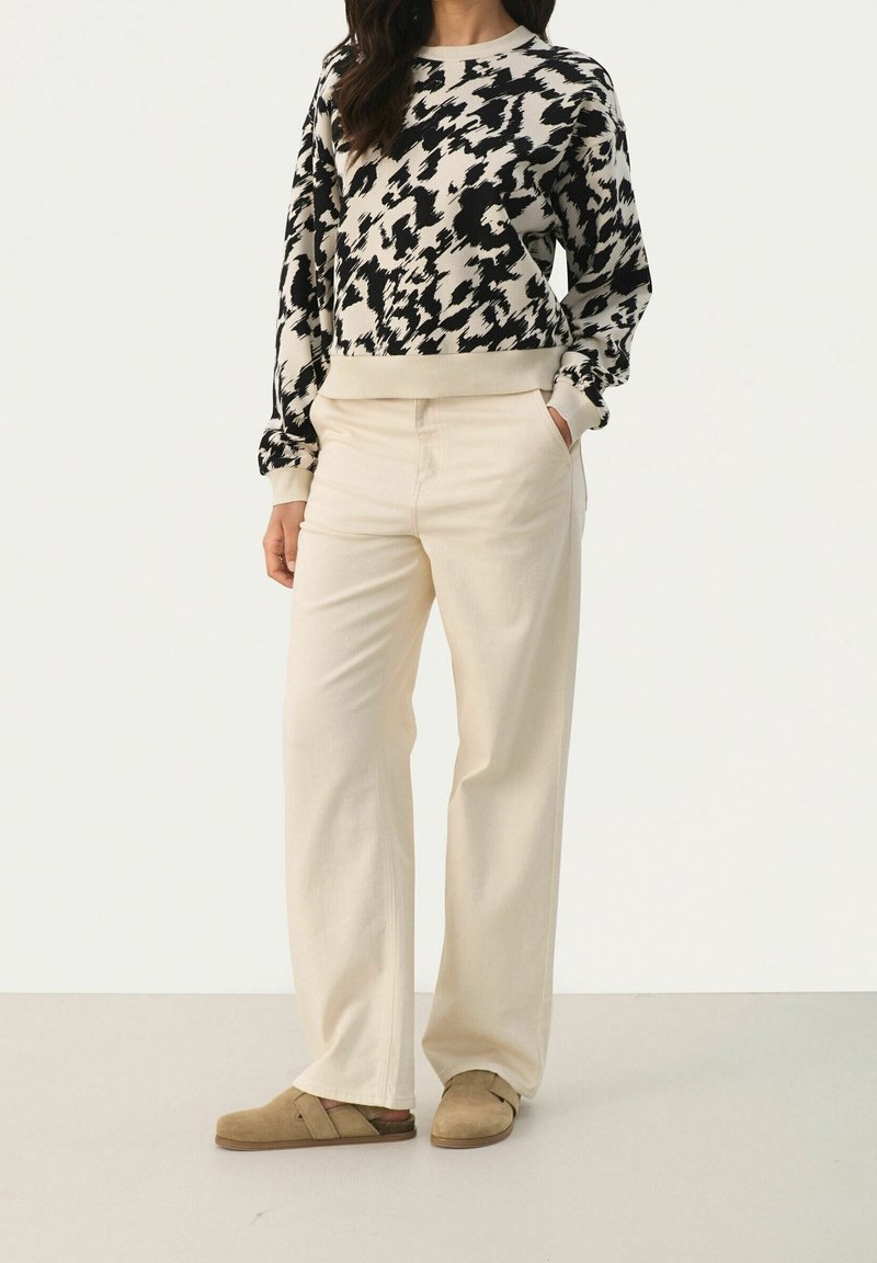 Femme portant un pantalon large beige, des chaussures beige à enfiler et un pull à manches longues avec un motif abstrait noir et blanc.