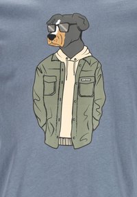 Graphic T-shirt featuring a Rottweiler in sunglasses, wearing a light hoodie and an olive green jacket with a patch, set on a blue background.